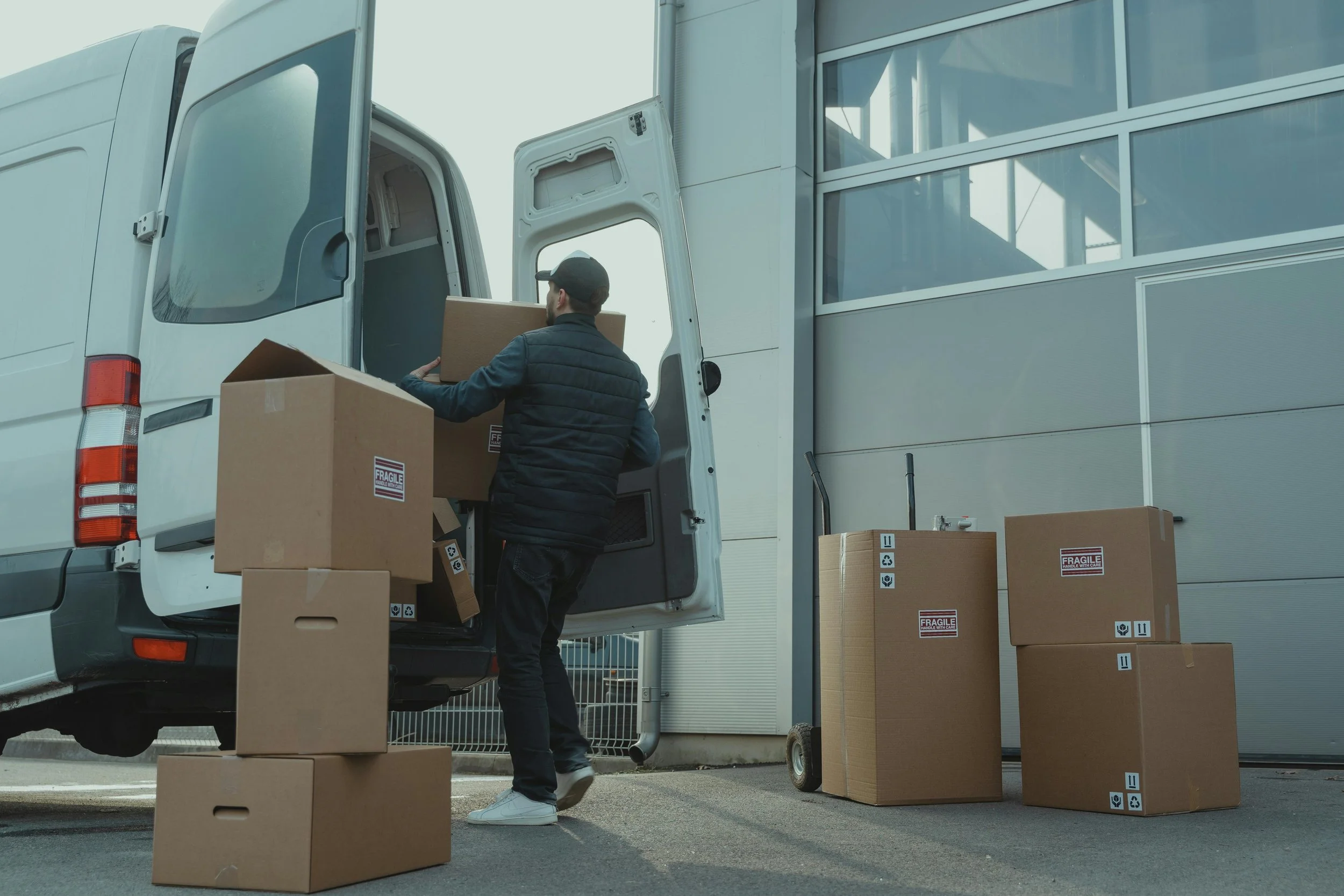 A man loading cardboard boxes into the back of a white delivery van outside a modern building, with more boxes on the ground nearby.