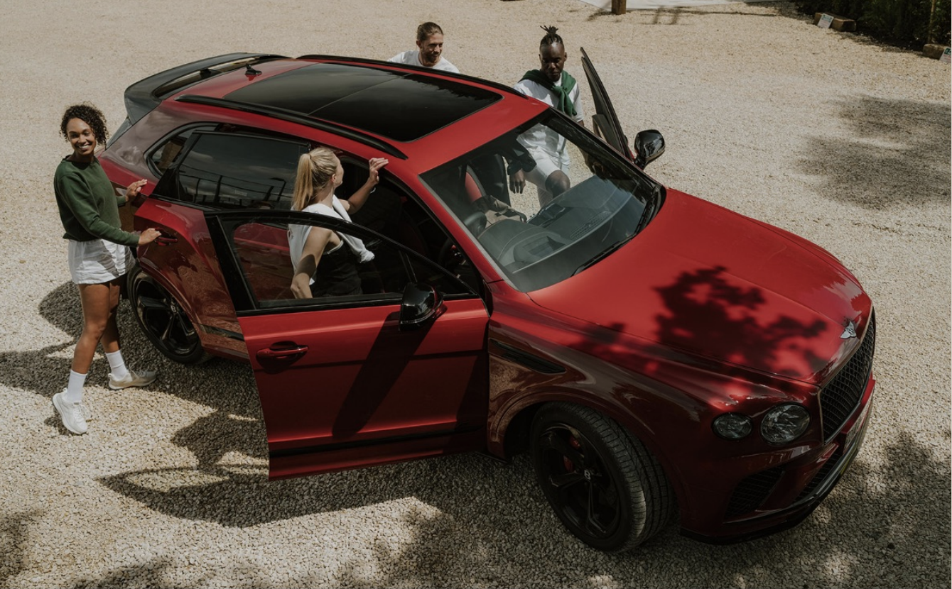 Four people standing around a red luxury car on a gravel surface, with two women standing outside, one woman inside the car, and a man near the open front passenger door.