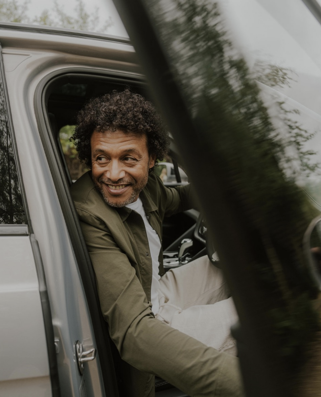 A man with curly hair smiling and sitting in the driver's seat of a vehicle, looking outside.