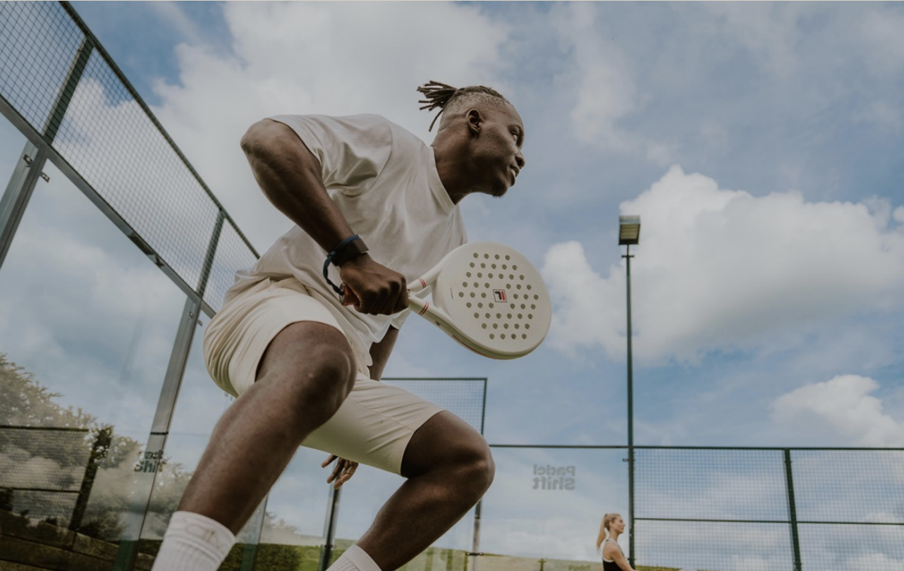 A man playing paddle tennis on an outdoor court, holding a paddle racket, wearing a white shirt and shorts, with a woman in the background near the court.