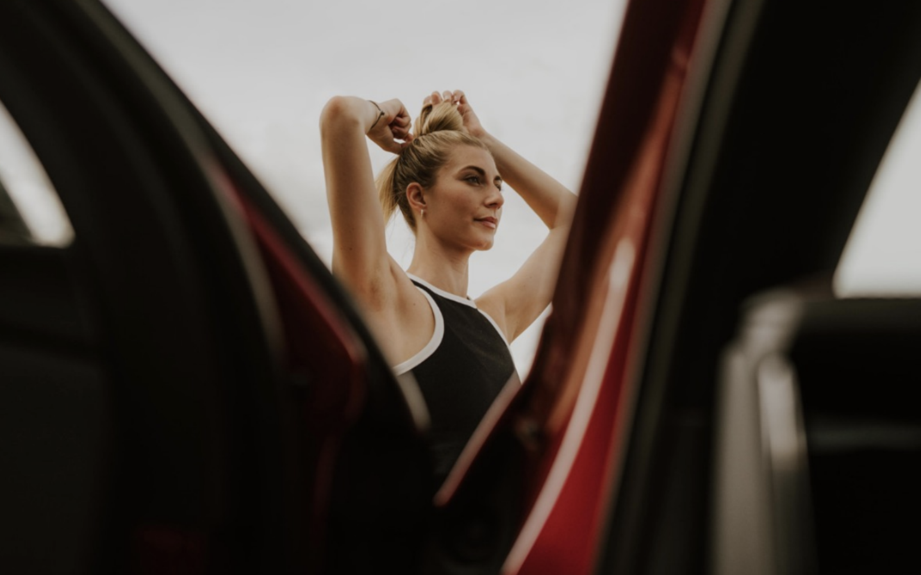 A woman standing outside, tying her hair into a bun, seen through the open door of a red car.