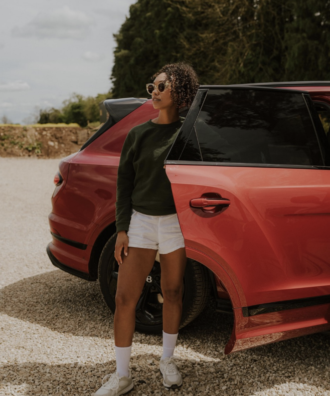 A woman standing next to a red SUV, wearing sunglasses, a dark green long-sleeve shirt, white shorts, white socks, and sneakers, outdoors on a gravel surface with trees in the background.