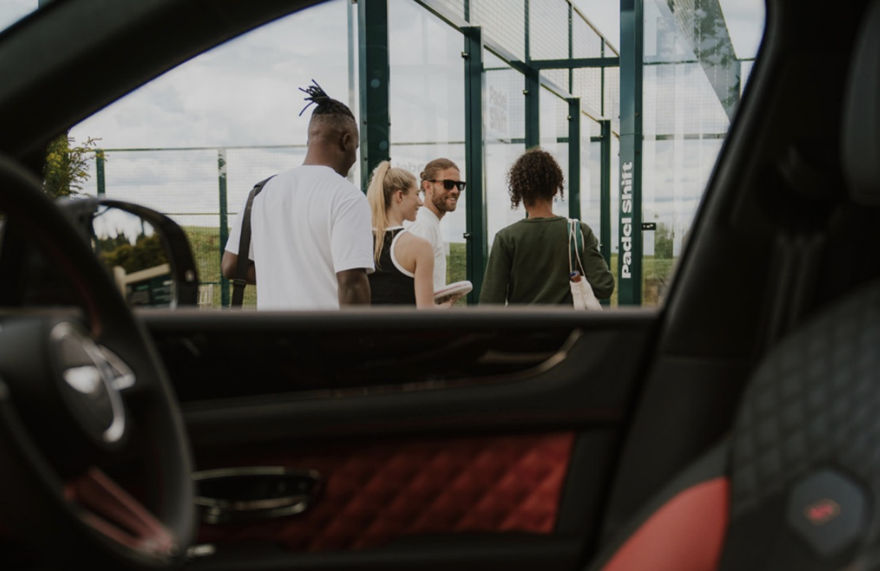 Four people standing at a padel tennis court entrance, seen through the car window, with a sign indicating 'Padel Shirt' on the fence.