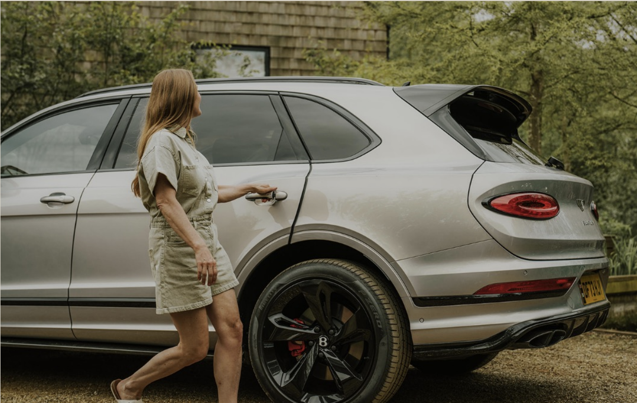 Woman standing next to a silver luxury SUV with a rear hatch open, outdoors surrounded by trees.