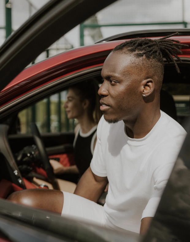 A man in a white t-shirt and white shorts sitting in a red car, with a woman in a black dress sitting in the driver's seat, smiling.