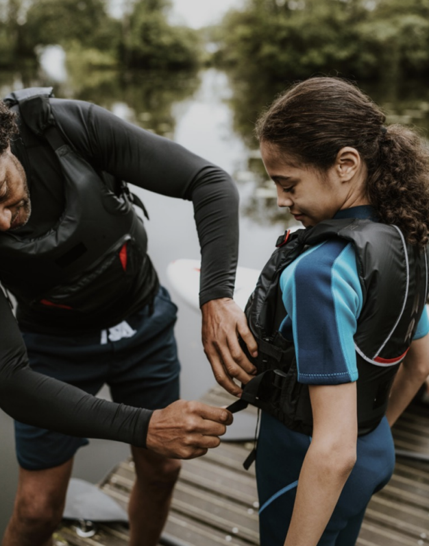 A man helps a young girl secure a life jacket before they go kayaking by a lake surrounded by trees.