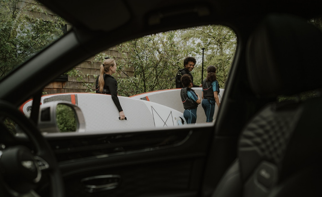 View of people holding surfboards and a group of young girls in wetsuits, seen through the car window on a cloudy day, with trees and a wooden wall in the background.
