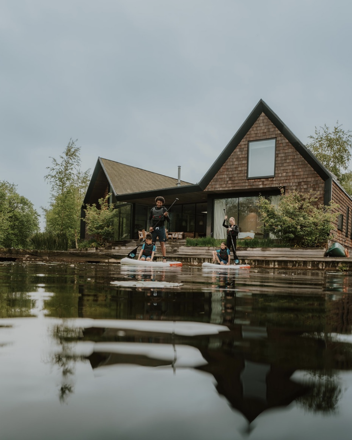 People paddleboarding on a lake in front of a modern house with a wooden exterior and large windows, surrounded by trees.