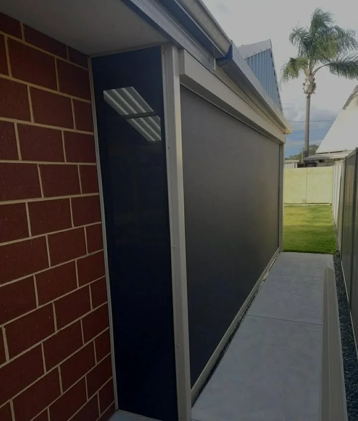 Side view of an outdoor screened-in porch or patio area with brick wall on the left, a metal fence on the right, and a grassy yard with palm trees in the background.