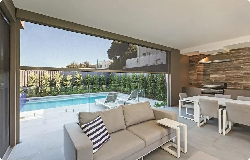 Modern living room with large glass sliding door opening to an outdoor pool area with lounge chairs, surrounded by greenery; interior features a beige sofa, striped pillow, and a dining table with six chairs; wooden accent wall with a mounted heater.