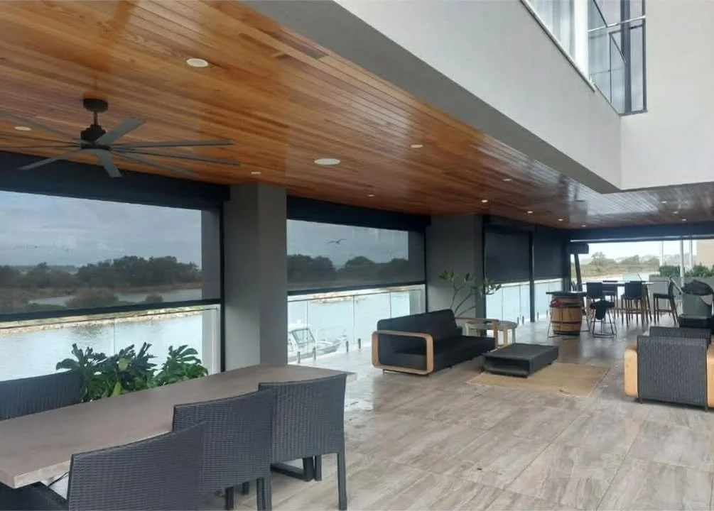 Interior of a modern waterfront lounge with large windows showing a river or lake view, featuring a wooden ceiling with a ceiling fan, various seating including a black couch, armchairs, and a dining table with chairs, and some indoor plants.