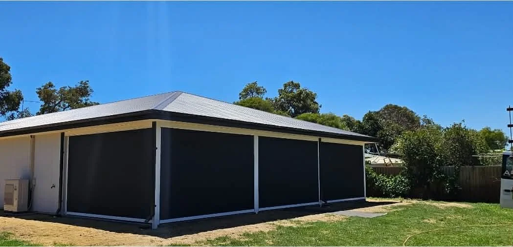 A black outdoor shed with a gray metal roof and white trim, located on a grassy area with trees in the background under a blue sky.