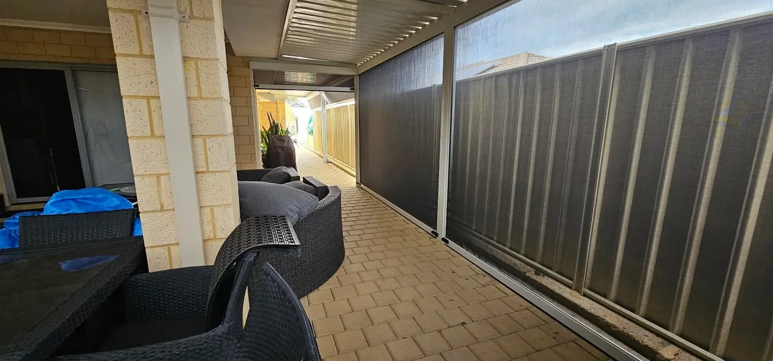 A covered outdoor patio area with brick flooring, wicker patio furniture with cushions, a dining table, and a screen enclosure along the side. There are plants and a yellow fence visible in the background.