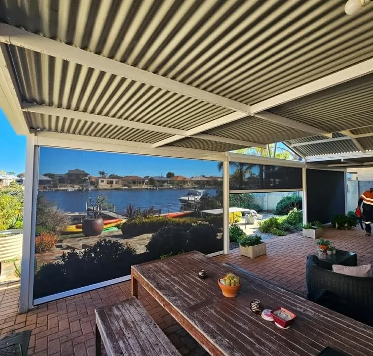 Outdoor patio with a trellis roof, overlooking a canal with houses and boats. There's a wooden table with potted plants and decorative items, and a person standing near the corner.