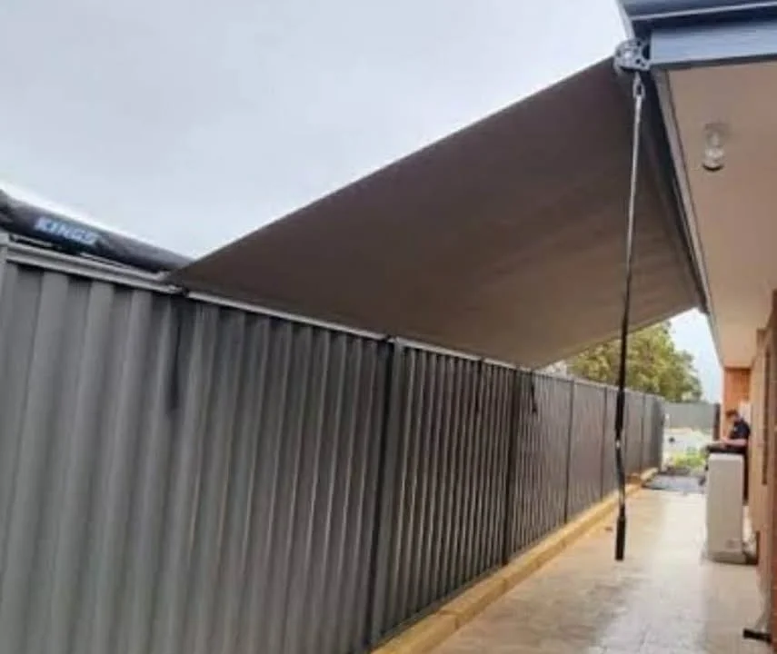 A large canopy or awning attached to a building extends outward, supported by a pole, with rain falling underneath it onto a sidewalk beside a fence.