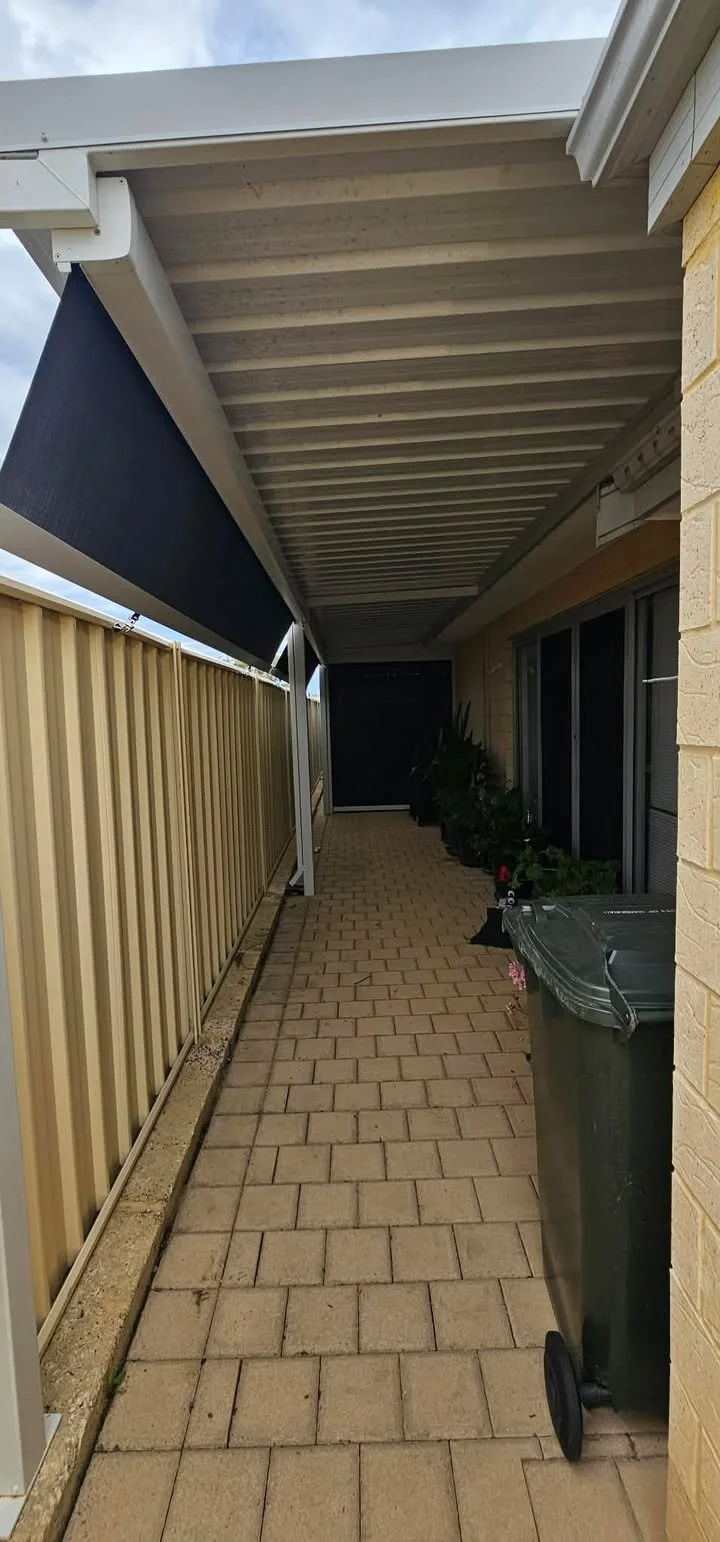 Narrow outdoor walkway with brick pavers, side fence, plants, trash bin, and awning overhead.