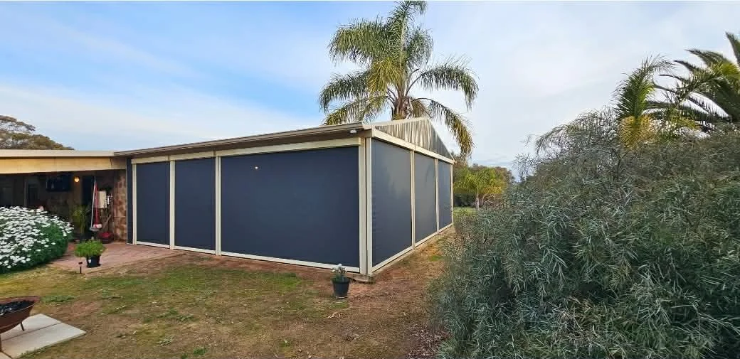 A house with an attached screened porch or patio, surrounded by plants and trees, including palm trees, in a yard with a mix of grass and dirt.