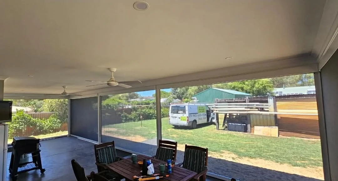Enclosed patio with a ceiling fan, a dining table with chairs and bottles or cans, a small grill, and a large view of outdoor yard with a van and sheds outside.