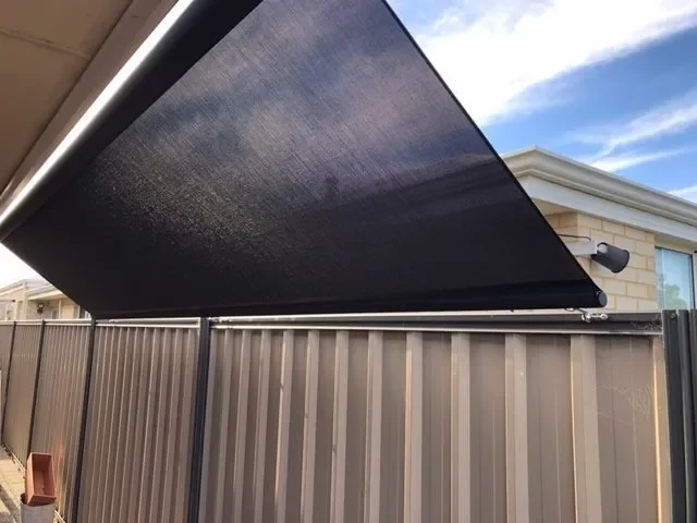 A black outdoor retractable awning extended over a fenced yard, with a house visible in the background and a partly cloudy sky.