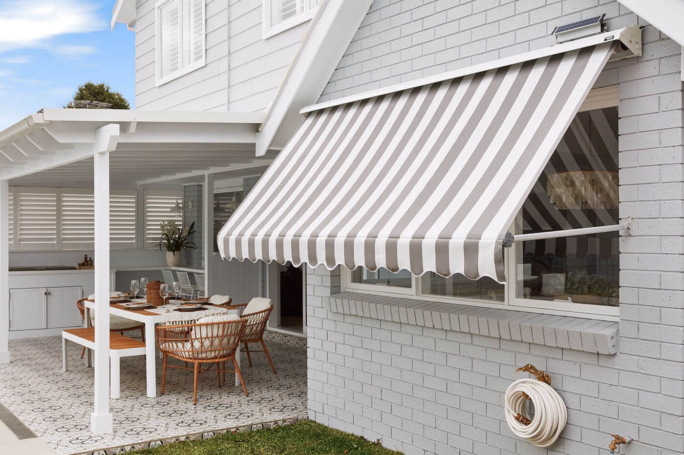 A house with a white brick exterior and a striped retractable awning over a window, with an outdoor dining area underneath a white pergola, featuring a wooden table, chairs, and tableware.