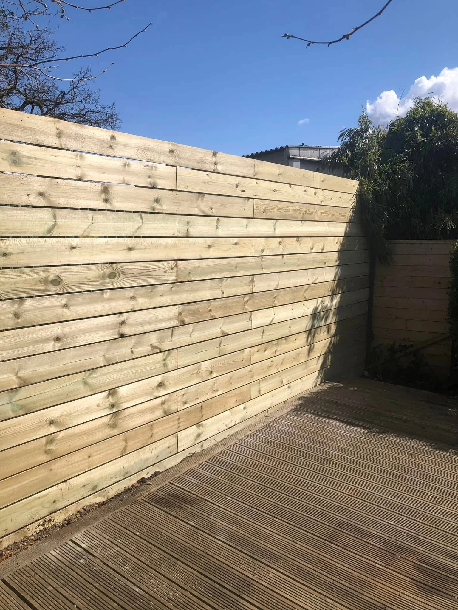 A newly constructed wooden fence dividing an outdoor space, with a wooden deck in the foreground, a tree with few leaves, and a clear blue sky with some clouds in the background.