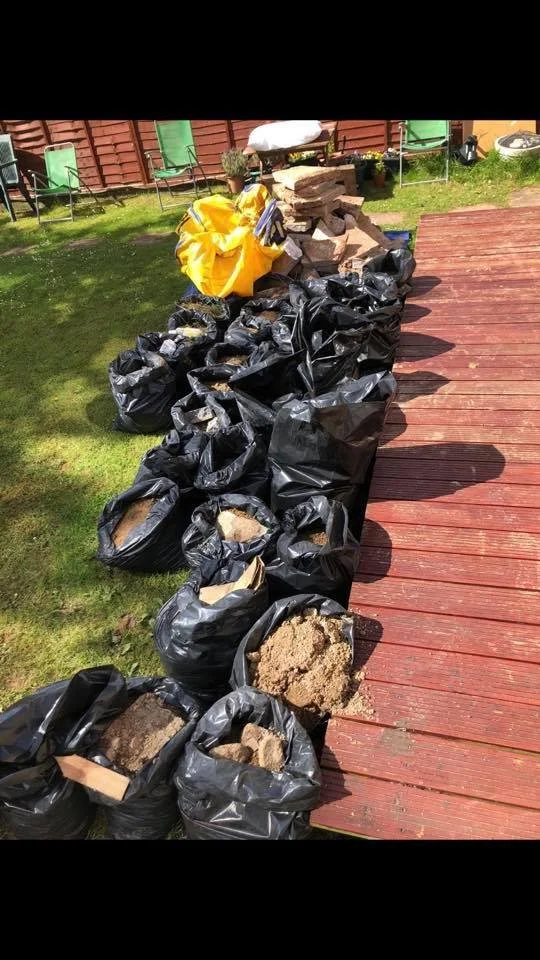 Black garbage bags filled with dirt and rocks lined up on a lawn next to a wooden deck, with some bricks and gardening supplies in the background.