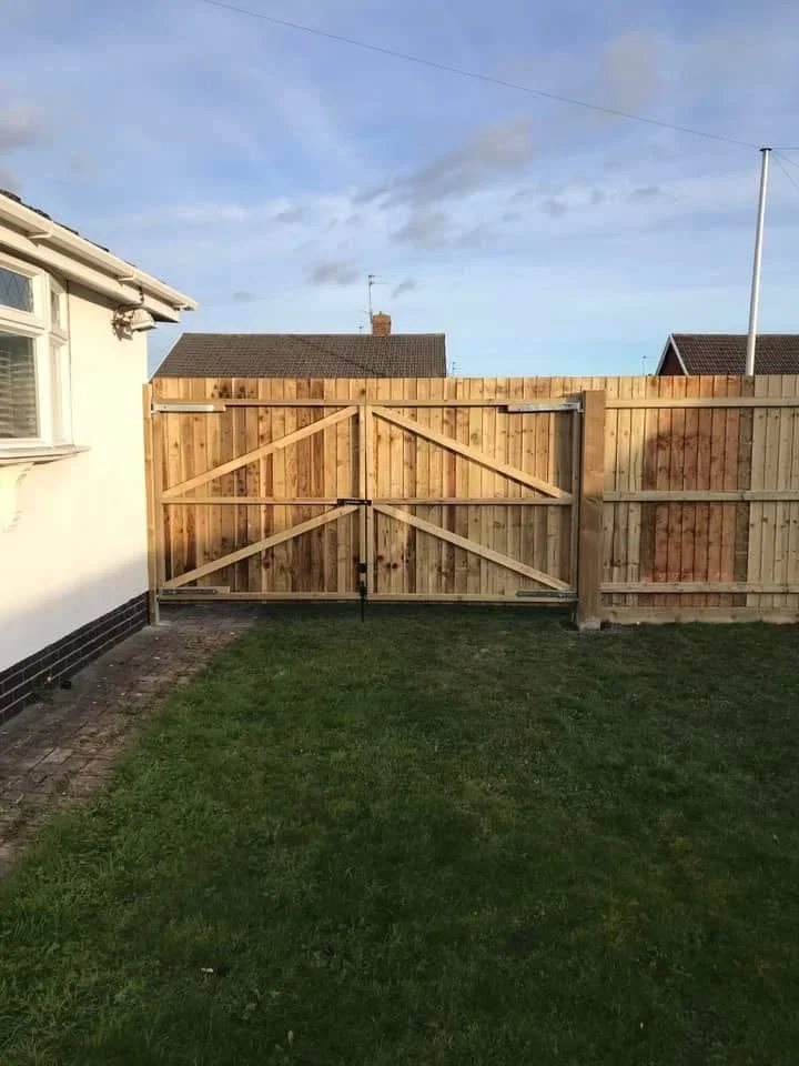 Backyard with a wooden gate, grass lawn, and neighboring houses in the background under a partly cloudy sky.