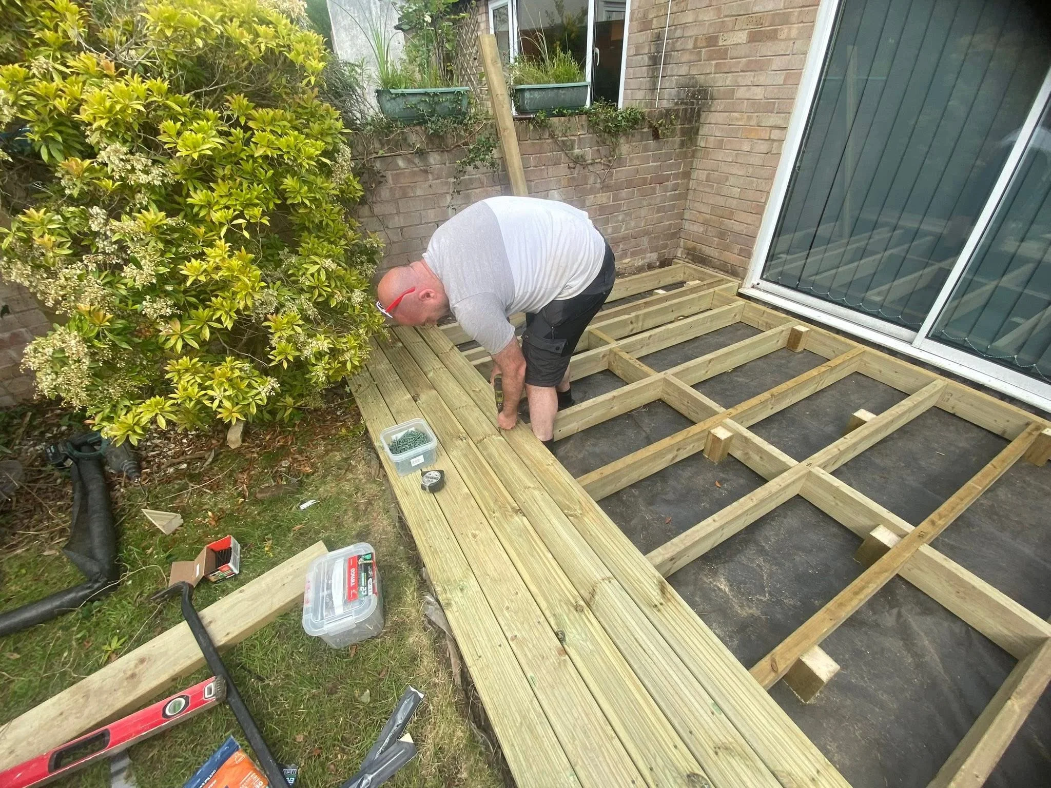 A man working on building a wooden deck outside a house, surrounded by tools and construction materials.