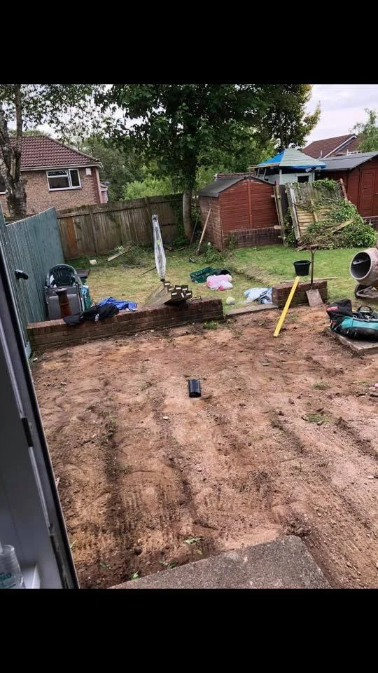 Backyard with partially excavated dirt area, construction tools, and materials, including a black pipe, yellow leveling tool, and small brick wall, with a fence, trees, and houses in the background.