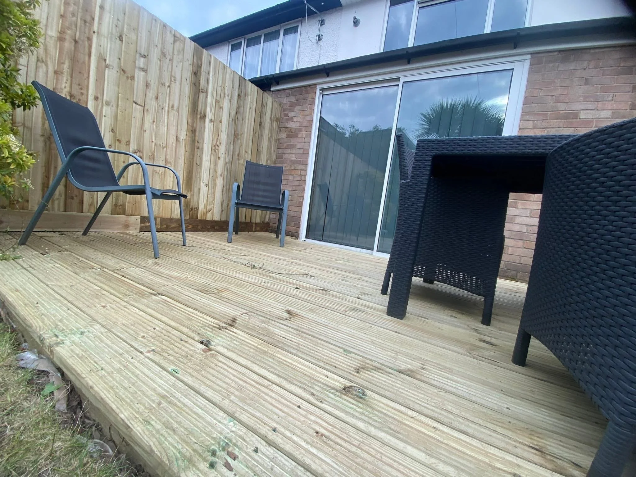 A backyard deck with outdoor furniture, including two black chairs, a black armchair, and a black wicker table, adjacent to a house with brick and siding exterior, and a sliding glass door.