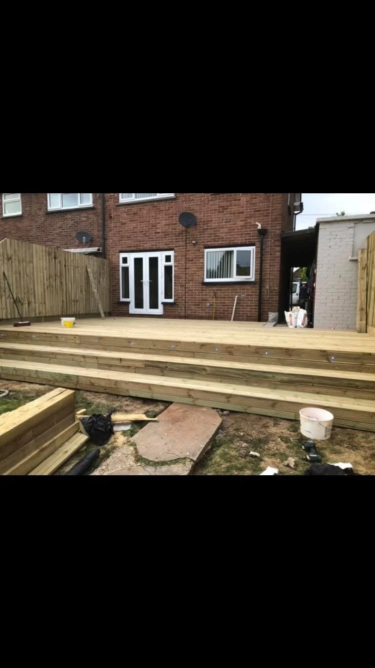 A backyard patio under construction with newly installed wooden decking, with building materials, tools, and a bucket visible in the area.
