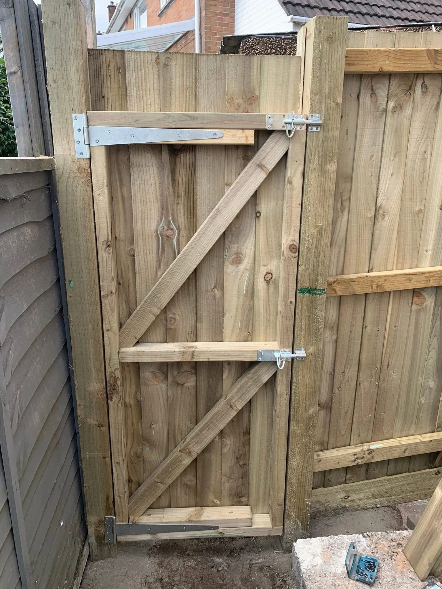 A newly installed wooden fence gate with metal hinges and latch, set in a backyard or garden area with neighboring fences and buildings in the background.