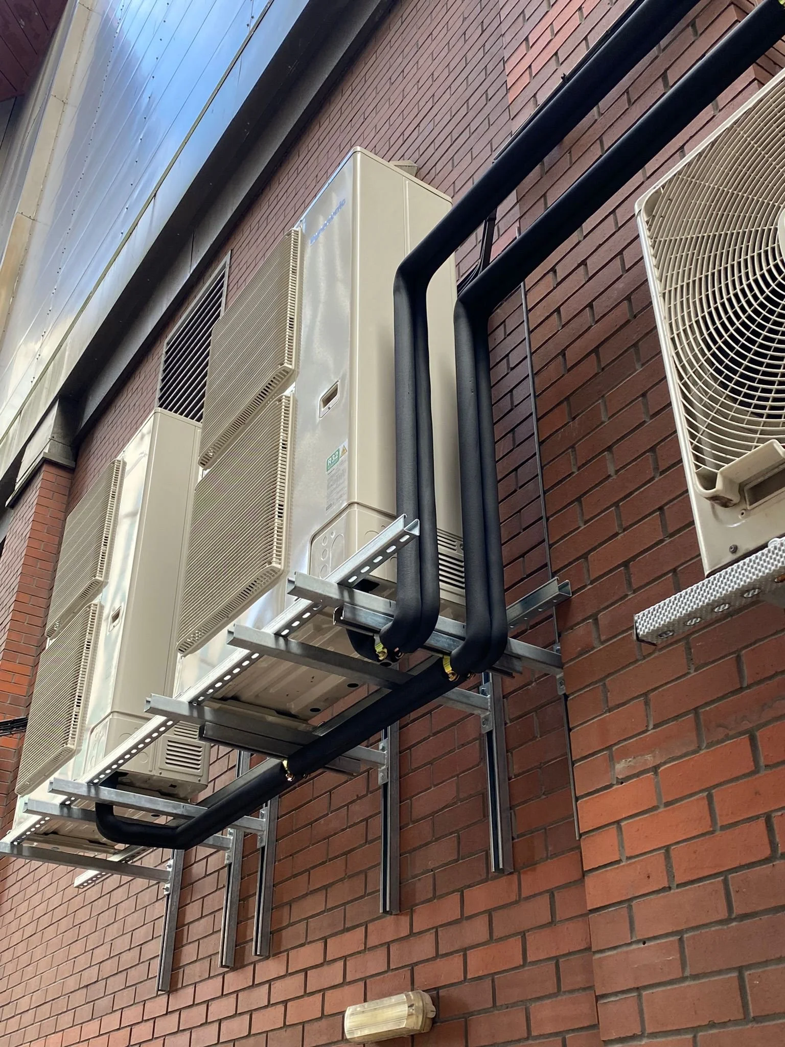 Three air conditioning units installed on a brick wall with black pipes and metal support brackets.