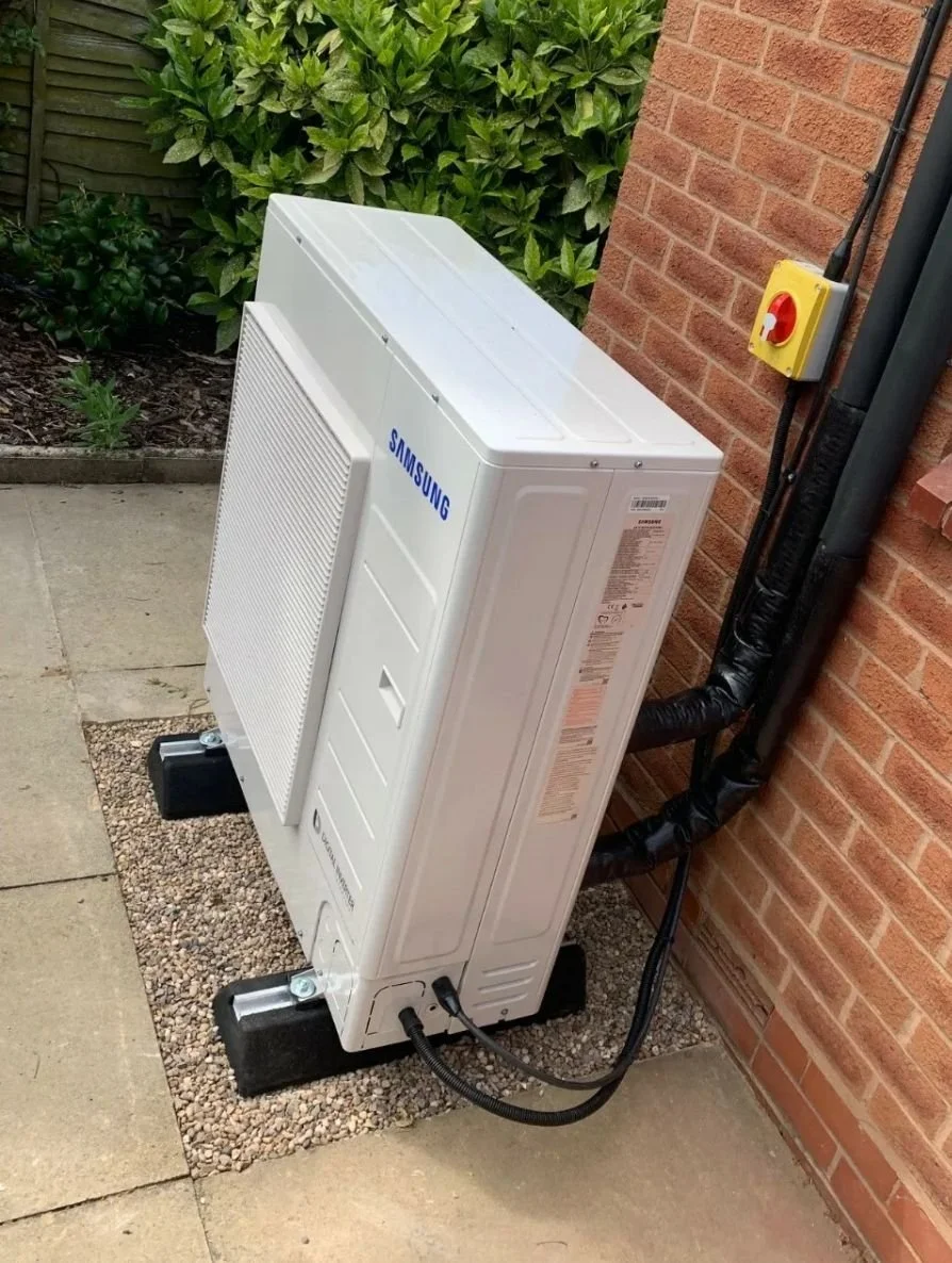 Outdoor Samsung heat pump or air conditioning unit installed next to a brick wall with electrical wiring and a red emergency stop button.