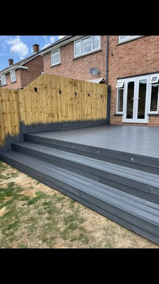 Garden deck with black outdoor flooring and wooden steps, enclosed by a wooden fence, attached to a brick house with glass doors.