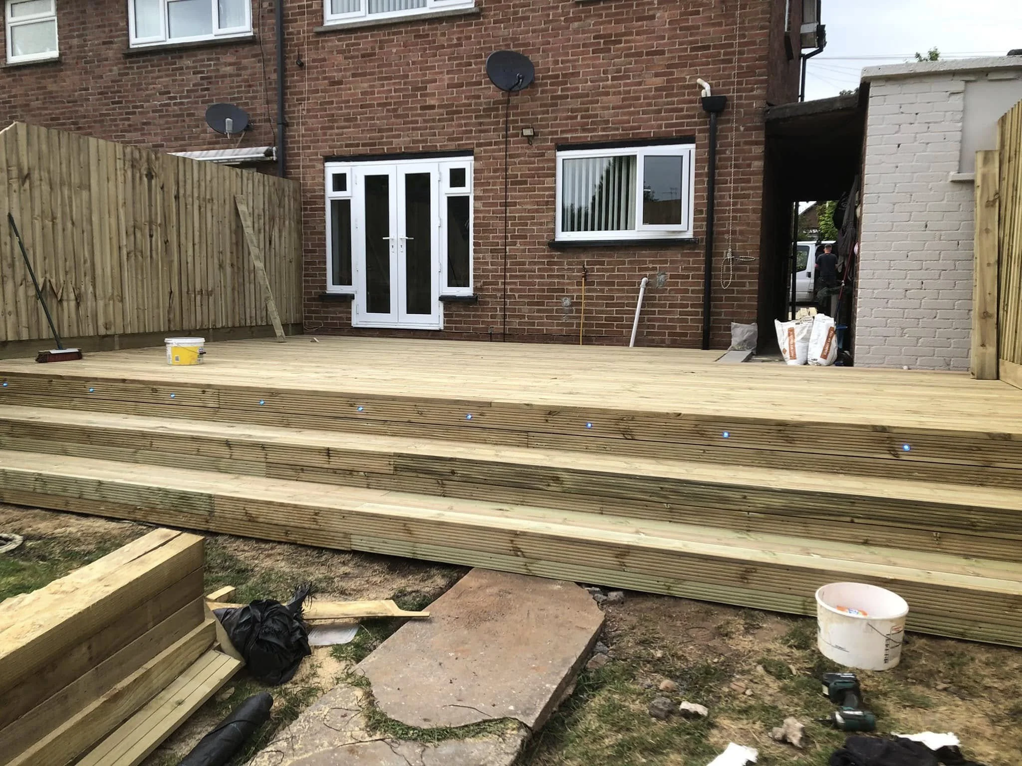 New wooden deck construction at the back of a brick house, with stairs leading up to it, construction tools, materials, and partially built sections visible.