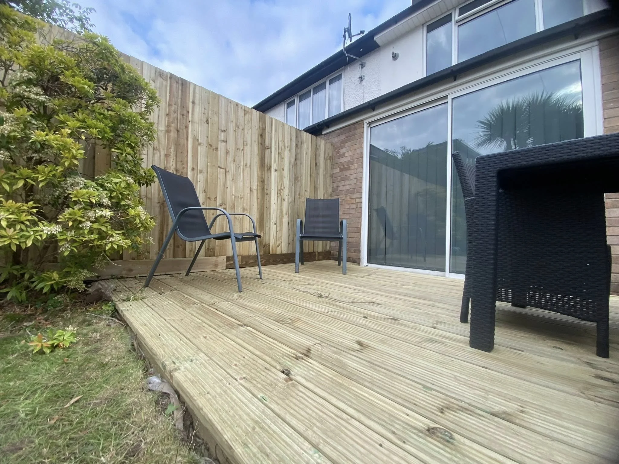 Backyard patio with wooden decking, two black chairs, a wooden fence, and a glass sliding door leading into a house.