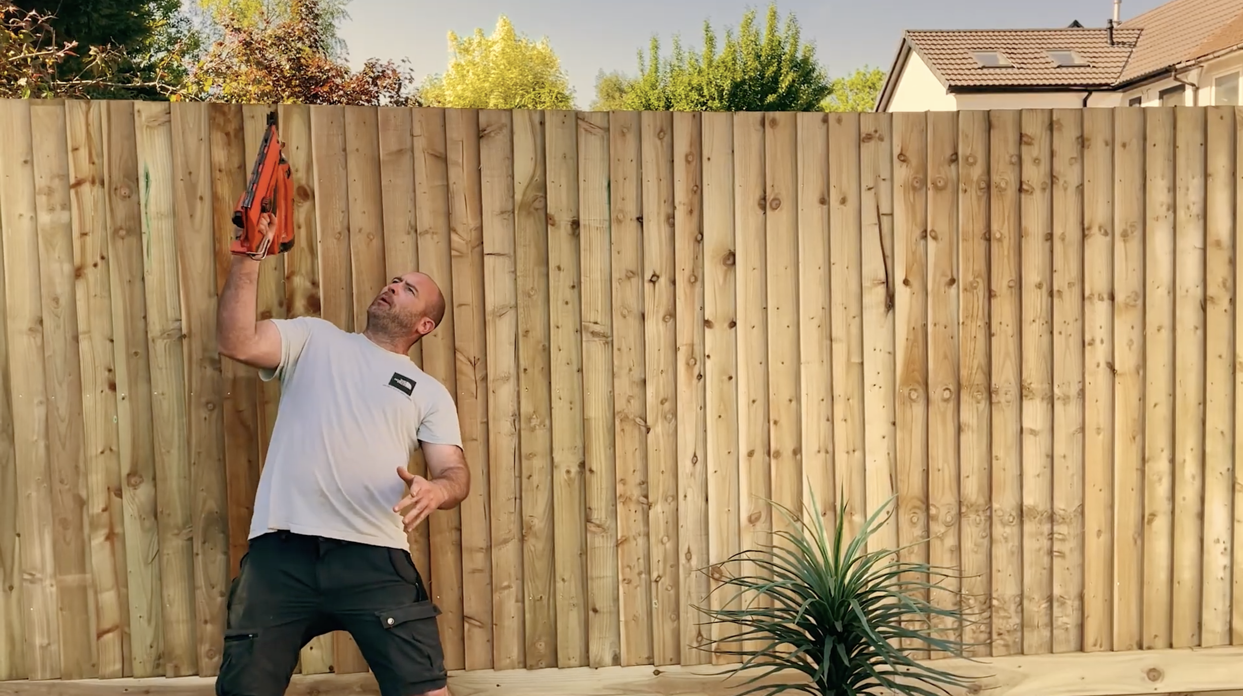 A man in a beige t-shirt and black shorts is holding and looking at an orange power tool, possibly a stapler or nail gun, outdoors in front of a wooden fence.