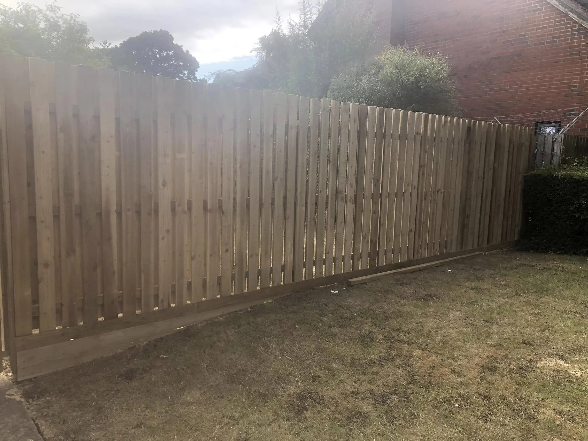 Newly installed wooden fence with vertical slats in a garden, bordered by grass and adjacent to a brick building.