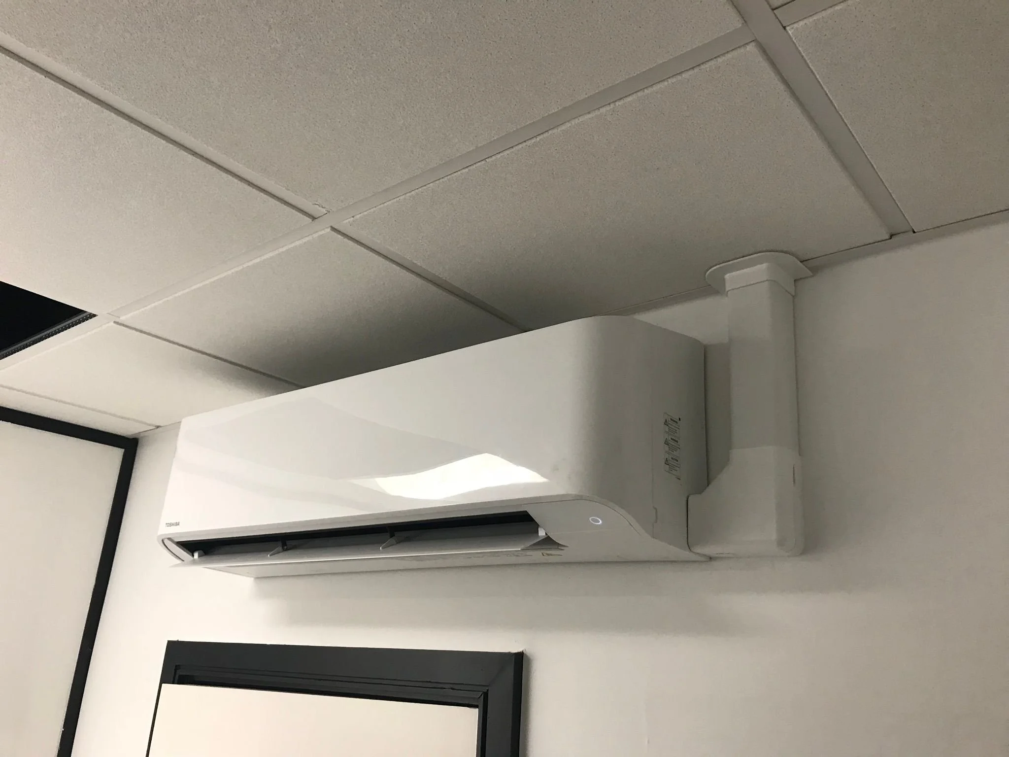 White wall-mounted air conditioner installed on a white wall, with a black border window below it.