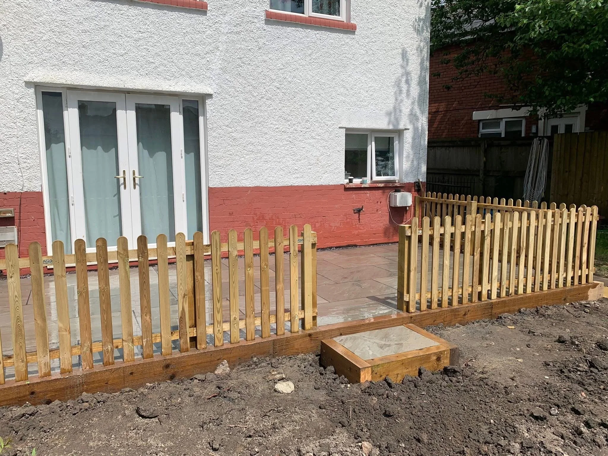 Backyard patio area with a wooden picket fence, a small raised platform, and a white house with sliding door and window.