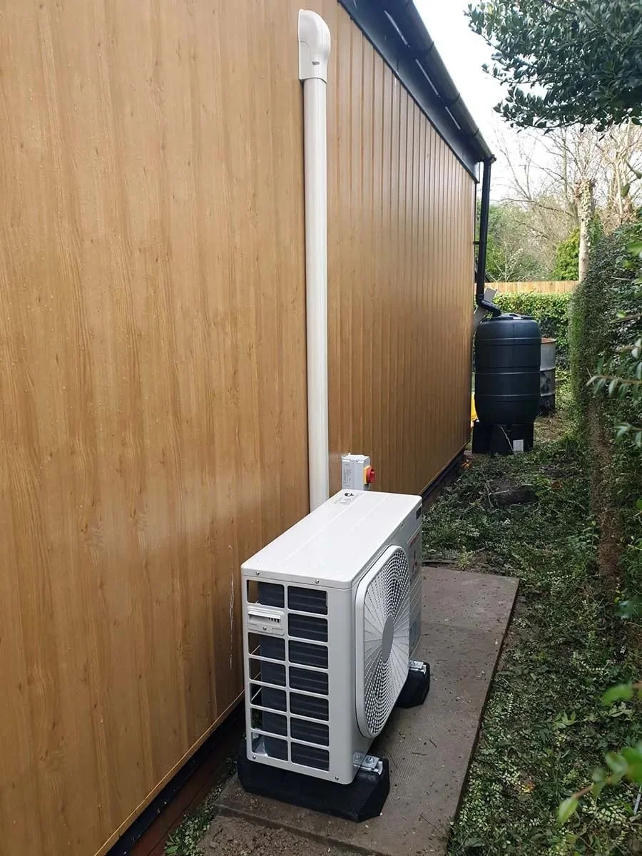 Outdoor air conditioning unit installed on a small concrete pad next to a wooden house wall, with a white drainpipe running down the wall and a black water barrel further in the background, surrounded by greenery.
