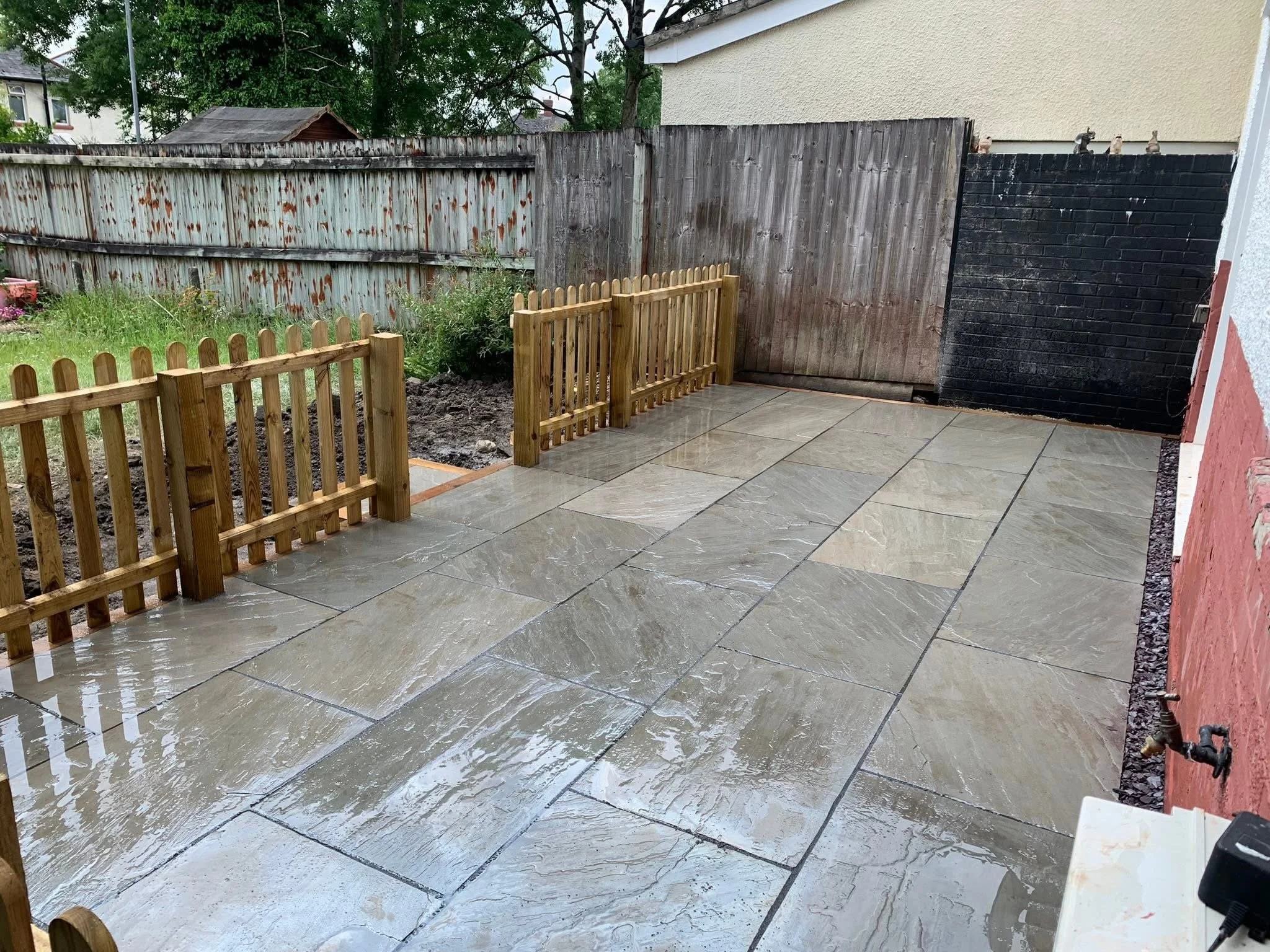 Wet outdoor patio with large stone tiles and a small wooden fence, next to a garden area with weeds and a weathered fence in the background.