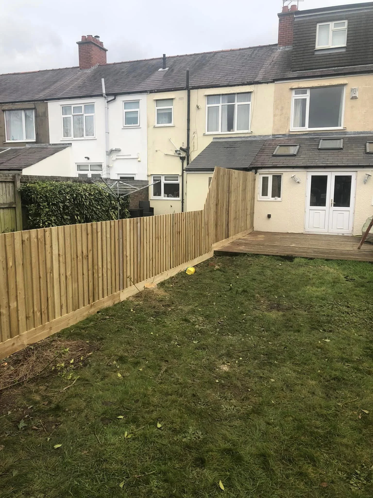 Backyard with a newly installed wooden fence, green grass, a wooden deck, and residential houses with windows and chimneys in the background.