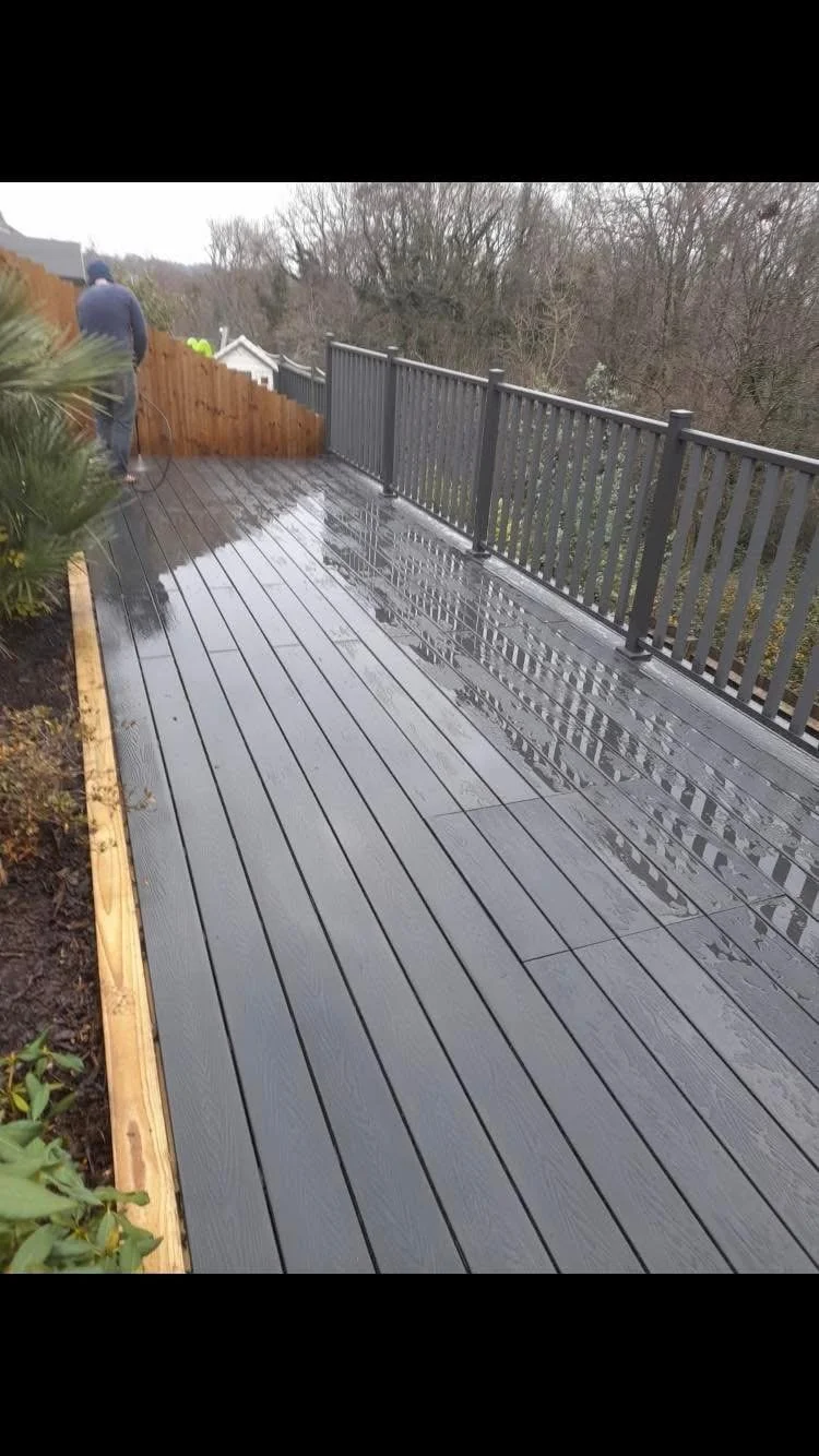 Wet wooden deck with a railing, next to a garden bed and a person walking away in the background.
