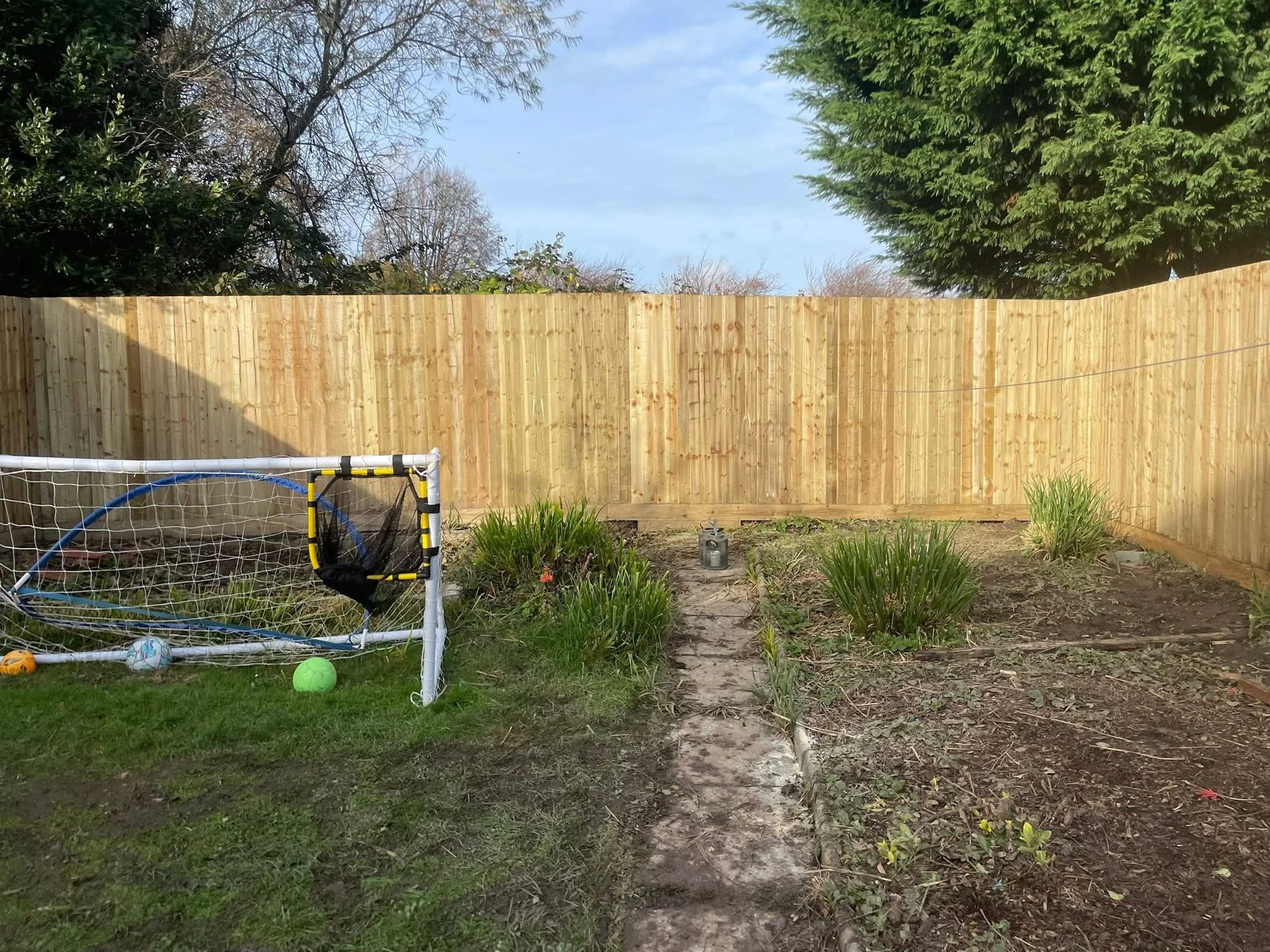 A backyard with a wooden fence, a small soccer goal with balls inside, and garden beds with green plants, trees, and a cloudy sky overhead.