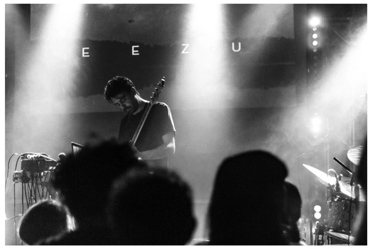 A black and white photo of Riccardo Tesorini playing a bass on stage with an electronic setup in front of him, and an audience in the foreground. The background has the letters 'EEZU' lit up.