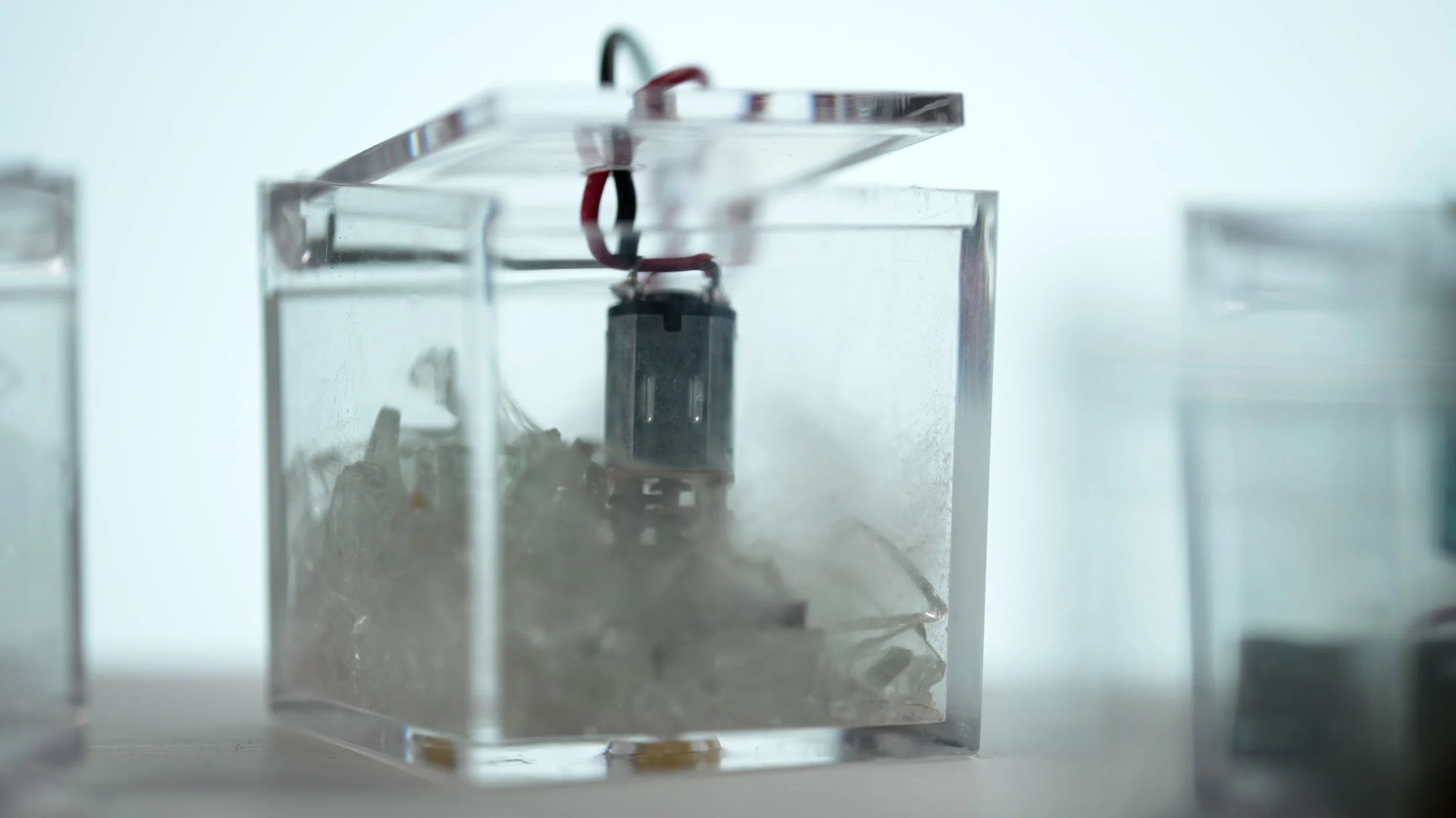 No one is an island Riccardo Tesorini sound installation Close-up of a small robotic device inside a transparent acrylic box filled with dust or debris, with wires attached to the top, and another similar box on the left and right sides.