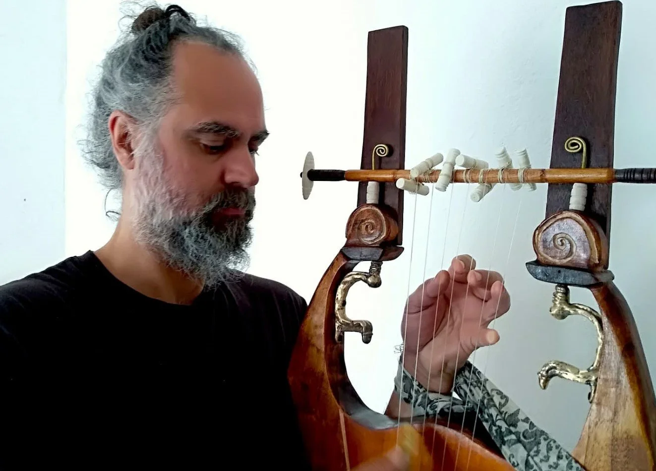 A man with gray hair and a beard, wearing a black shirt, playing a wooden harp with white strings, set against a plain white wall.