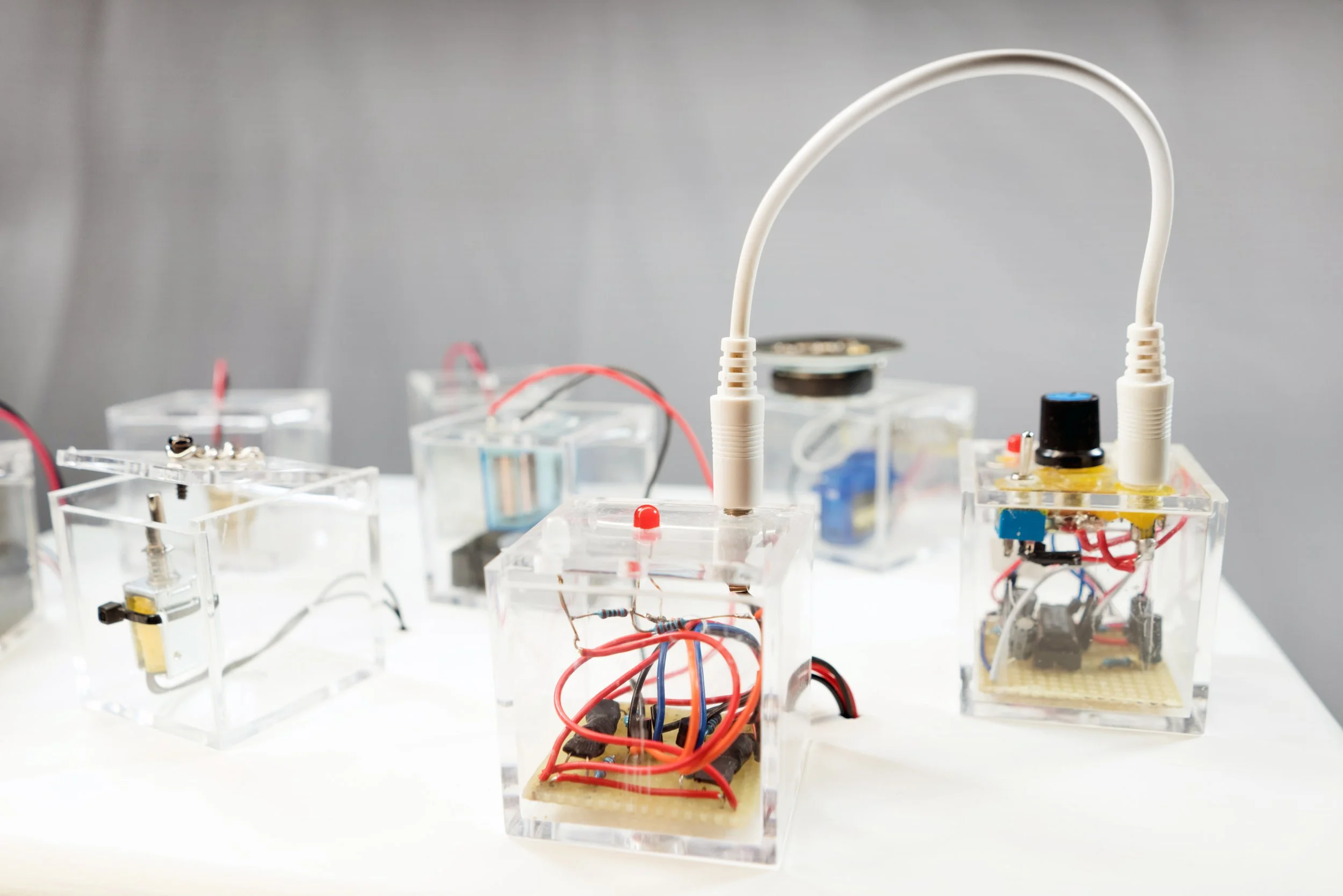 No one is an island Riccardo Tesorini  Multiple small electronic devices encased in transparent blocks with visible wiring and circuitry, placed on a white surface against a plain background. sound installation